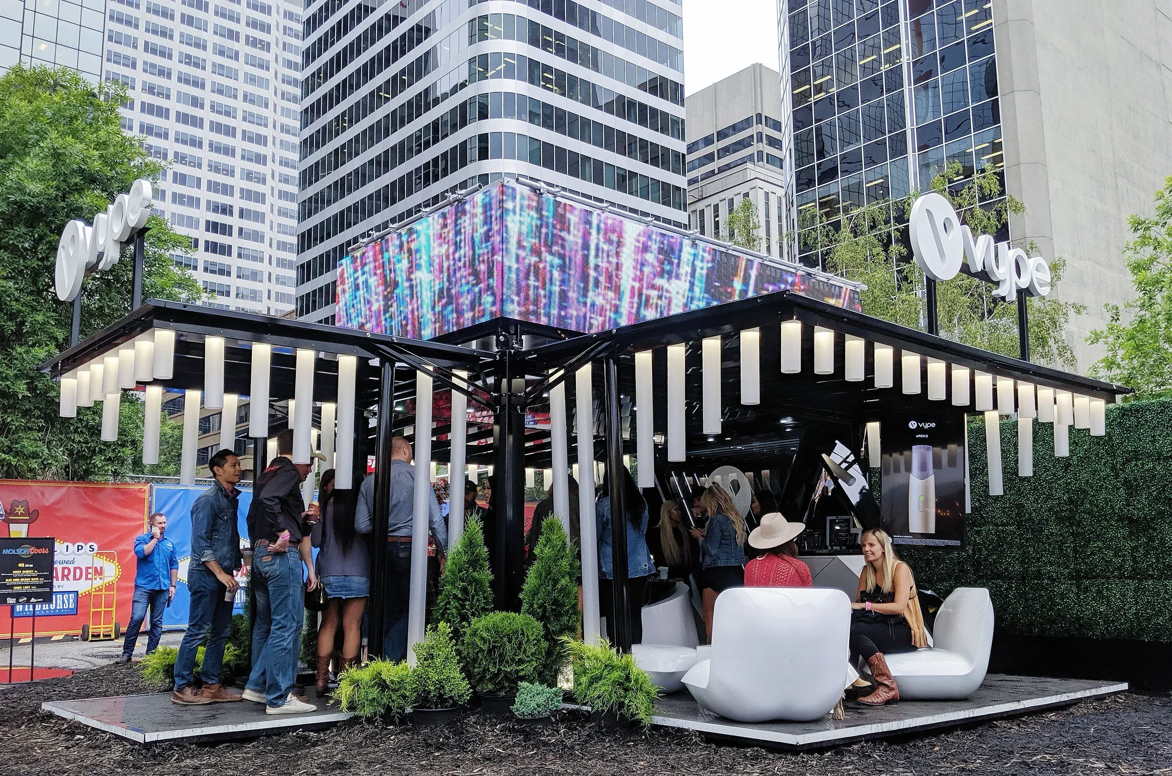 The Vype festival pavilion by day in a downtown plaza, its black steel canopy and glowing baton lights over white lounge seating, planting, and a digital media wall, with guests gathered among surrounding towers