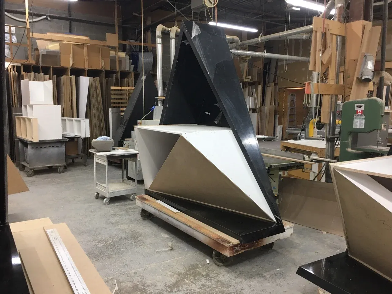 A faceted, tapered pavilion piece on a rolling dolly in the fabrication shop, its black outer shell open to reveal white and raw MDF inner panels, surrounded by a band saw, work tables, and stacked sheet stock