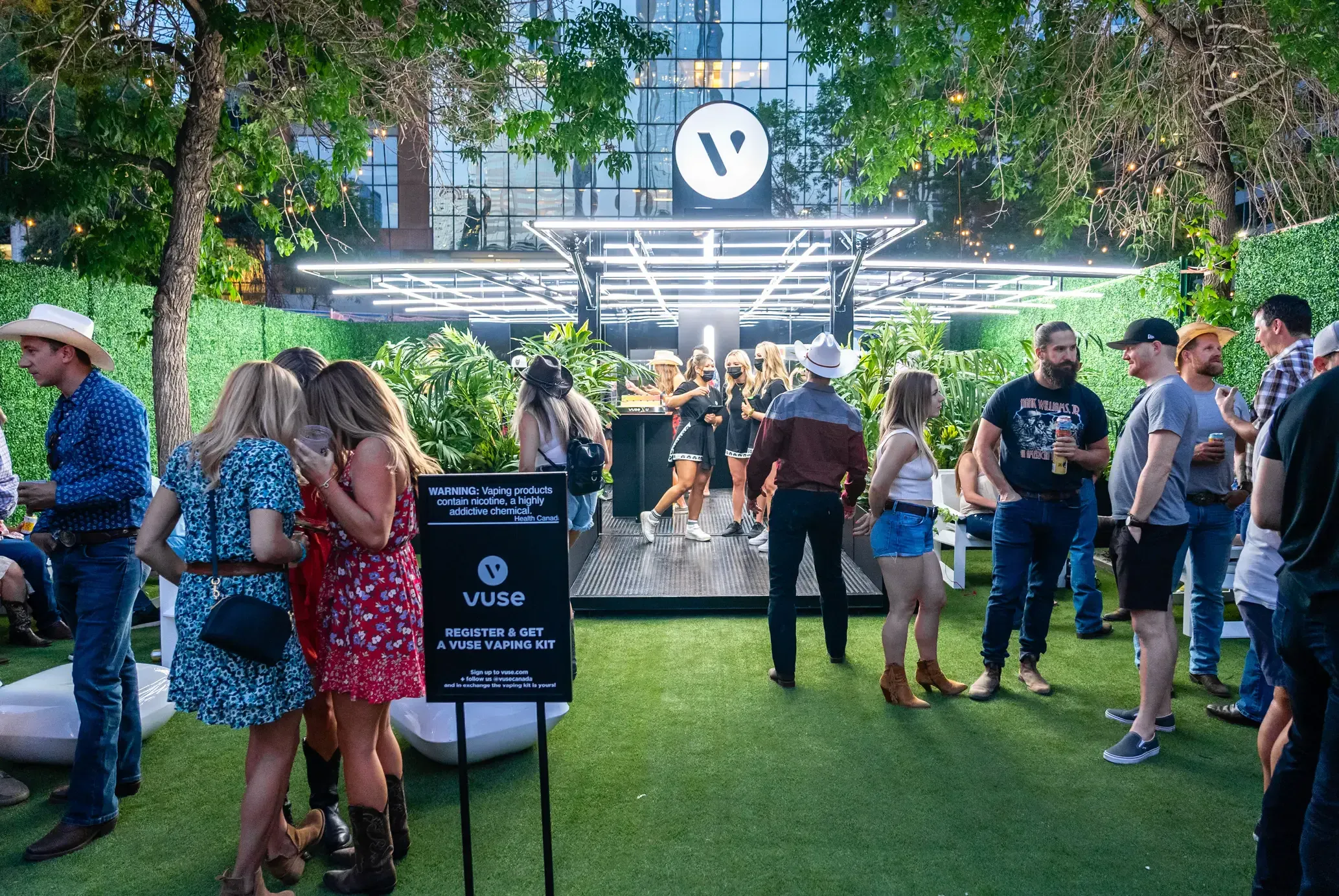 The rebranded Vuse festival activation outdoors at dusk, its lit canopy and rooftop Vuse roundel over a hedged lounge, with a cowboy-hatted crowd gathered around a registration sign on the grass