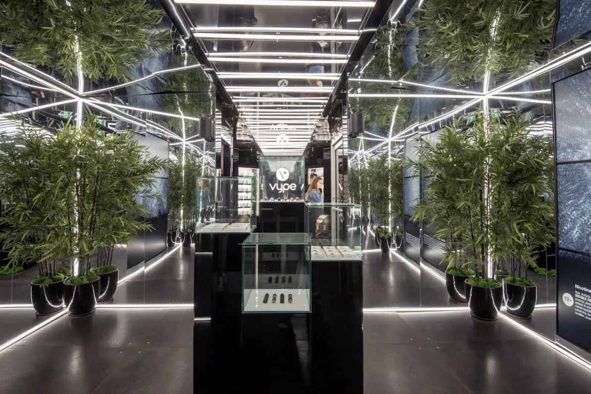 The mirrored interior of the Vuse container at night, walls and ceiling lined with white light strips that reflect into infinity, potted bamboo down both sides and glass product cases around a central Vuse-branded counter