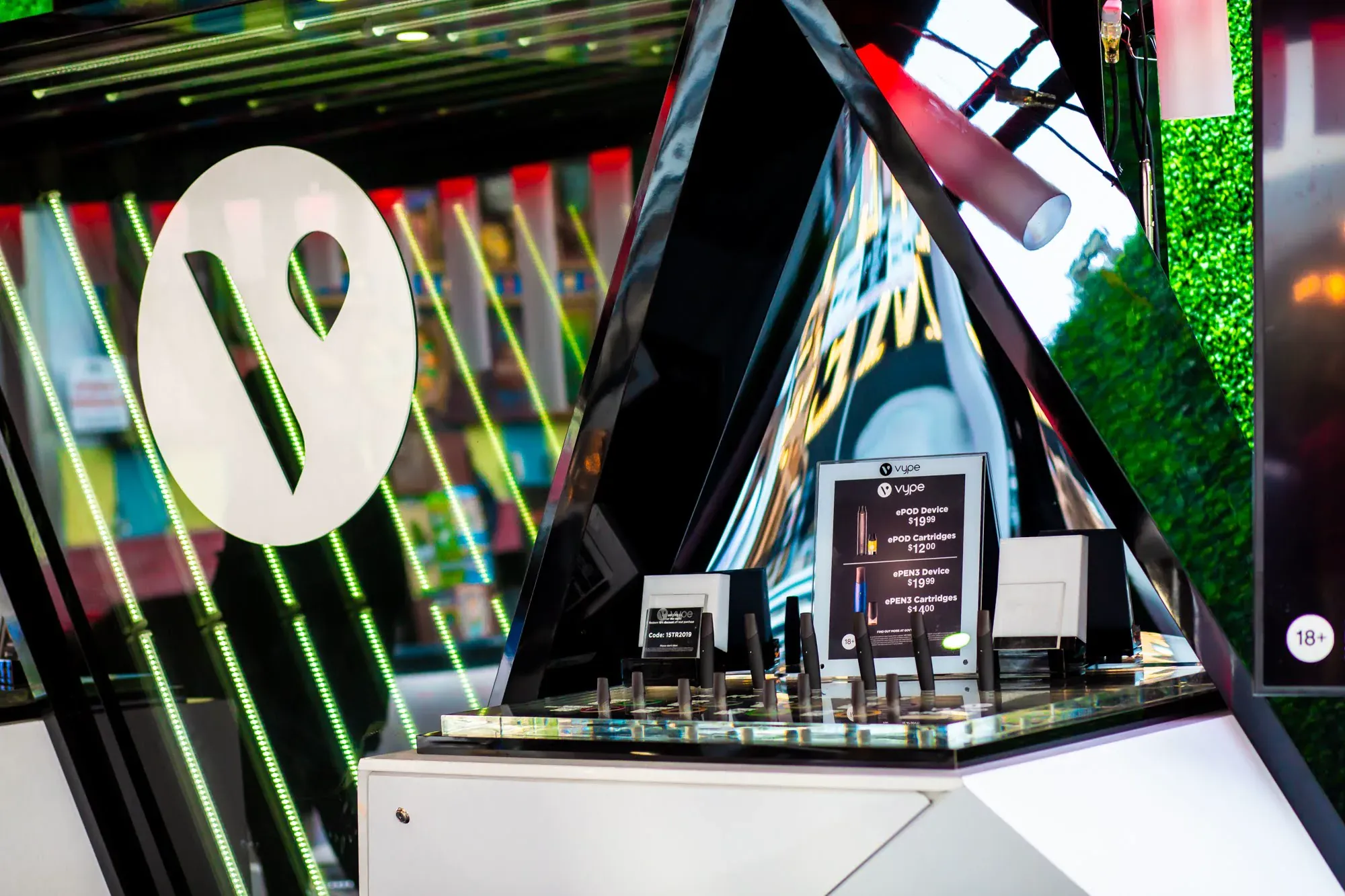 Close-up of the mirrored, triangular Vype product display with the white Vype logo cut into a backlit green-lit panel, devices and ePOD cartridges arranged on the glass counter with a price card