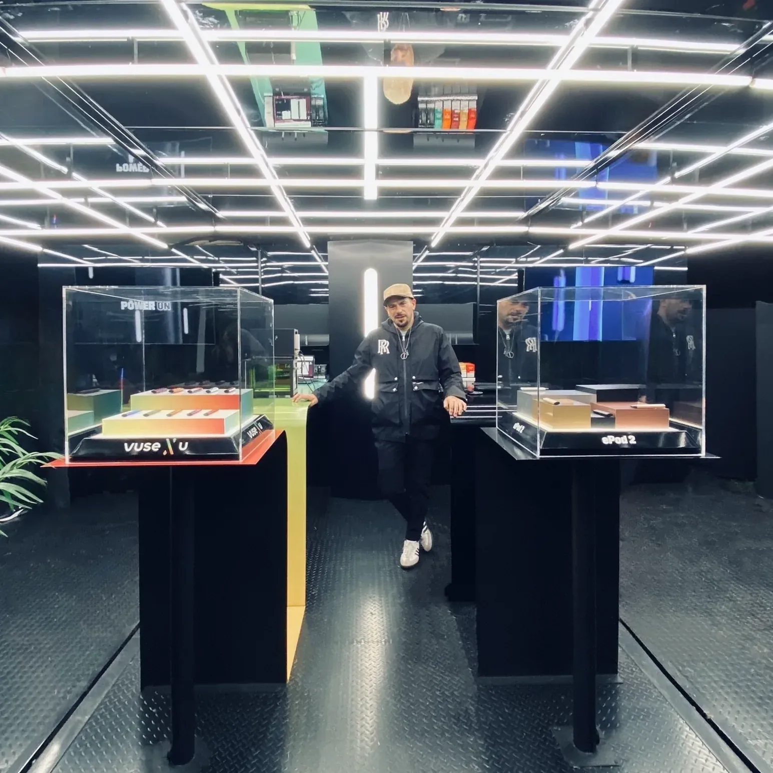 The interior of the container activation at night, lit by a dense overhead grid of white light bars reflected in mirrored ceilings, with two illuminated acrylic display cases presenting the Vuse devices and pods on a dark, reflective floor