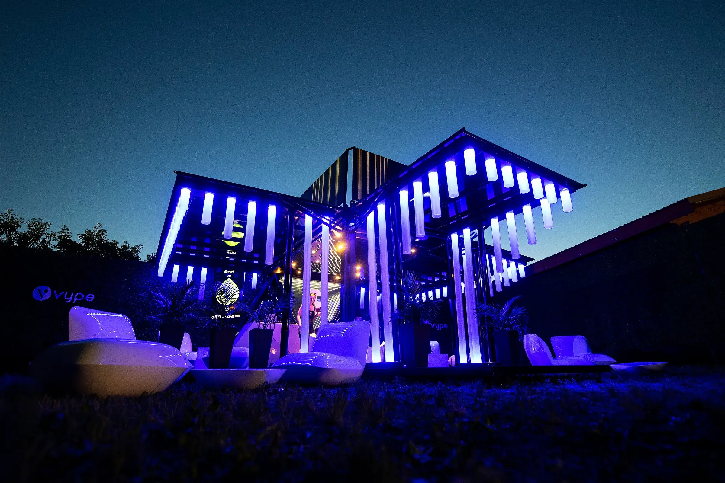 The same pavilion at night, its structural batons lit deep blue and white so the canopy reads as a glowing landmark across the festival field, with illuminated lounge furniture below
